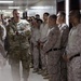 A U.S. Army officer in Operational Camouflage Pattern (OCP) uniform stands with arms crossed, appearing to address or brief a large group of military personnel in a hallway. The assembled group includes personnel wearing both OCP and Marine Corps MARPAT desert camouflage uniforms, suggesting a joint-service gathering. The setting appears to be an indoor military facility corridor.