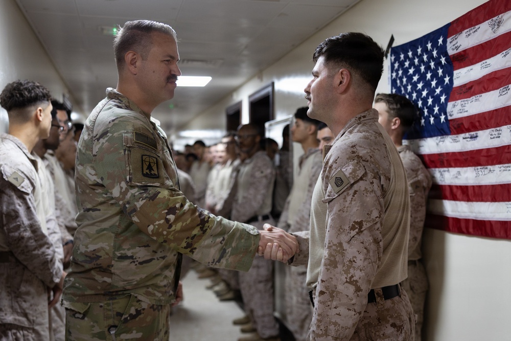 A U.S. Army soldier in OCP (Operational Camouflage Pattern) uniform is shaking hands with a U.S. Marine in MARPAT desert uniform in an indoor corridor. Multiple service members are lined up along the wall in the background, with a signed American flag displayed prominently. The setting appears to be a military installation, possibly during a formal visit or recognition ceremony.