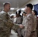 A U.S. Army soldier in OCP (Operational Camouflage Pattern) uniform is shaking hands with a U.S. Marine in MARPAT desert uniform in an indoor corridor. Multiple service members are lined up along the wall in the background, with a signed American flag displayed prominently. The setting appears to be a military installation, possibly during a formal visit or recognition ceremony.