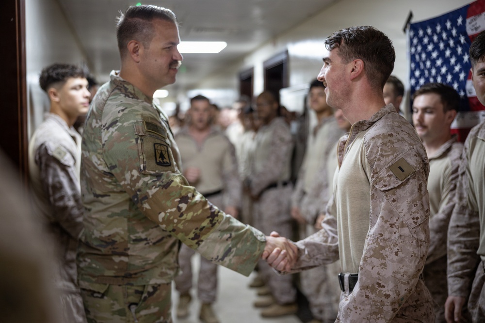 A U.S. Army soldier in OCP (Operational Camouflage Pattern) uniform is shaking hands with a U.S. Marine in MARPAT desert camouflage uniform in what appears to be an indoor military facility. A group of additional military personnel in desert camouflage uniforms lines the hallway in the background. An American flag is visible on the wall to the right.