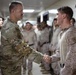 A U.S. Army soldier in OCP (Operational Camouflage Pattern) uniform is shaking hands with a U.S. Marine in MARPAT desert camouflage uniform in what appears to be an indoor military facility. A group of additional military personnel in desert camouflage uniforms lines the hallway in the background. An American flag is visible on the wall to the right.