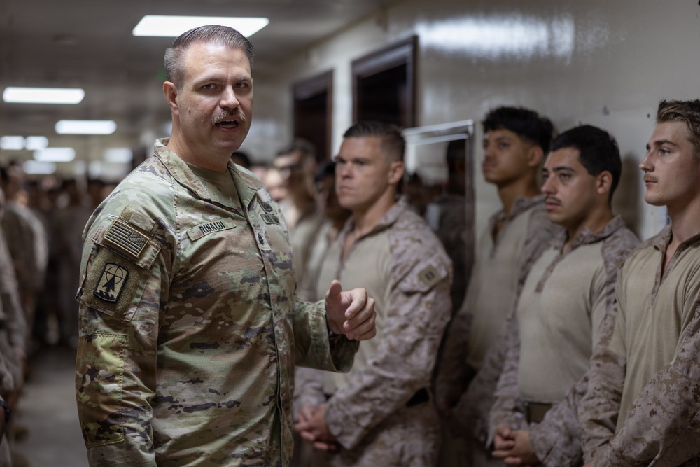 A U.S. Army soldier with the name tape 'RINALDI' and wearing OCP (Operational Camouflage Pattern) uniform is addressing a group of U.S. Marines in MARPAT desert uniforms inside what appears to be a military building or barracks hallway. The Army soldier appears to be a senior NCO or officer speaking to the assembled Marines. The setting suggests a briefing, inspection, or command visit.