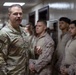 A U.S. Army soldier with the name tape 'RINALDI' and wearing OCP (Operational Camouflage Pattern) uniform is addressing a group of U.S. Marines in MARPAT desert uniforms inside what appears to be a military building or barracks hallway. The Army soldier appears to be a senior NCO or officer speaking to the assembled Marines. The setting suggests a briefing, inspection, or command visit.