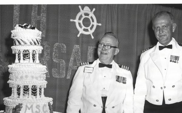 Two men in white naval or maritime dress uniforms with bow ties and medals stand beside a large multi-tiered celebration cake. The cake is decorated with the initials 'M.S.C.' and leaf motifs. A ship's wheel decoration and partial text reading 'RSARY' (likely 'ANNIVERSARY') are visible on the backdrop behind them.