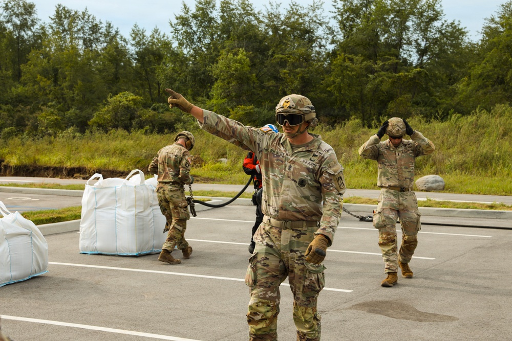 Three U.S. Army soldiers in OCP (Operational Camouflage Pattern) uniforms are conducting what appears to be a sling load or helicopter external lift operation in a paved area. The central soldier is giving a hand signal with one arm extended, wearing protective goggles and gloves, while holding a cable or static line. Large white bulk cargo bags are visible on the left side of the frame.