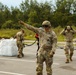 Three U.S. Army soldiers in OCP (Operational Camouflage Pattern) uniforms are conducting what appears to be a sling load or helicopter external lift operation in a paved area. The central soldier is giving a hand signal with one arm extended, wearing protective goggles and gloves, while holding a cable or static line. Large white bulk cargo bags are visible on the left side of the frame.