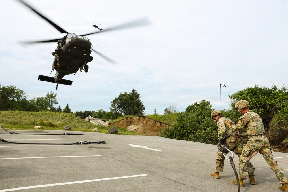 Two soldiers in U.S. Army OCP (Operational Camouflage Pattern) uniforms and helmets are handling a fuel hose or sling load hook on a paved surface while a UH-60 Black Hawk helicopter hovers at low altitude nearby. The scene appears to depict a forward area refueling point (FARP) or sling load training exercise. Ropes and rigging equipment are visible on the ground around them.