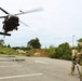 Two soldiers in U.S. Army OCP (Operational Camouflage Pattern) uniforms and helmets are handling a fuel hose or sling load hook on a paved surface while a UH-60 Black Hawk helicopter hovers at low altitude nearby. The scene appears to depict a forward area refueling point (FARP) or sling load training exercise. Ropes and rigging equipment are visible on the ground around them.