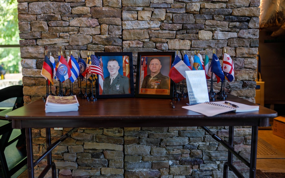 A memorial or ceremonial display table set against a stone wall, featuring two framed military portrait photographs of uniformed Marines flanked by numerous small international and U.S. state/organizational flags. The table also holds printed materials, a sign-in notebook with a pen, and what appears to be a stack of programs or pamphlets. The arrangement suggests a formal military event such as a retirement ceremony, memorial service, or change of command.