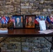 A memorial or ceremonial display table set against a stone wall, featuring two framed military portrait photographs of uniformed Marines flanked by numerous small international and U.S. state/organizational flags. The table also holds printed materials, a sign-in notebook with a pen, and what appears to be a stack of programs or pamphlets. The arrangement suggests a formal military event such as a retirement ceremony, memorial service, or change of command.