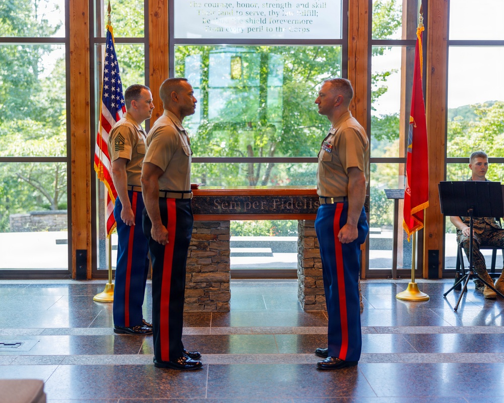 Three U.S. Marines in dress uniform stand at attention inside a formal venue featuring a 'Semper Fidelis' engraved podium or altar. Two Marines face each other in what appears to be a military ceremony, while a third stands at parade rest behind them. An American flag and a Marine Corps flag flank the scene, with a musician visible in the background.