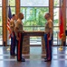 Three U.S. Marines in dress uniform stand at attention inside a formal venue featuring a 'Semper Fidelis' engraved podium or altar. Two Marines face each other in what appears to be a military ceremony, while a third stands at parade rest behind them. An American flag and a Marine Corps flag flank the scene, with a musician visible in the background.