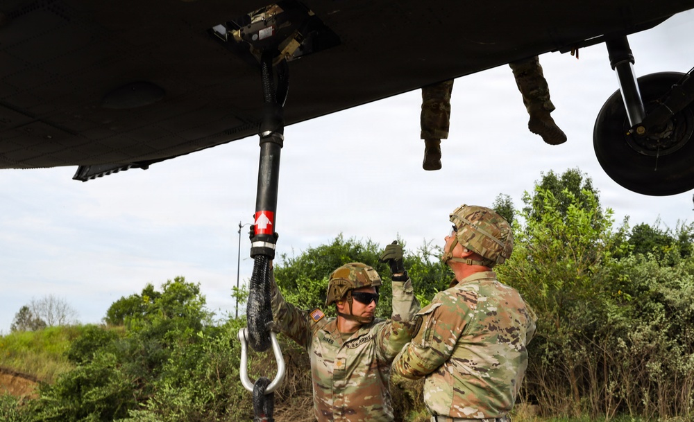 Two U.S. Army soldiers in OCP (Operational Camouflage Pattern) uniforms are working beneath a military helicopter, handling what appears to be a cargo hook or sling load attachment system. A third soldier is visible from the waist down standing on top of or inside the aircraft. The scene appears to be a field training exercise involving helicopter sling load operations.