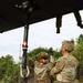 Two U.S. Army soldiers in OCP (Operational Camouflage Pattern) uniforms are working beneath a military helicopter, handling what appears to be a cargo hook or sling load attachment system. A third soldier is visible from the waist down standing on top of or inside the aircraft. The scene appears to be a field training exercise involving helicopter sling load operations.