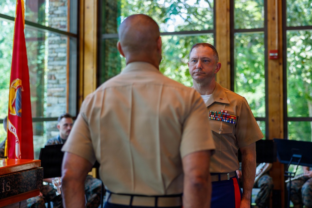 Two U.S. Marine Corps officers in tan service uniforms (Charlie uniform) face each other in what appears to be a formal military ceremony. The officer facing the camera displays multiple rows of service ribbons on his chest. The setting appears to be an indoor venue with large windows overlooking greenery, with a Marine Corps flag visible in the background.