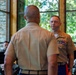 Two U.S. Marine Corps officers in tan service uniforms (Charlie uniform) face each other in what appears to be a formal military ceremony. The officer facing the camera displays multiple rows of service ribbons on his chest. The setting appears to be an indoor venue with large windows overlooking greenery, with a Marine Corps flag visible in the background.