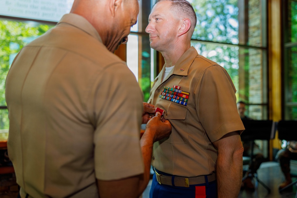 Two U.S. Marine Corps personnel in tan service uniform (Charlie uniform) are engaged in a military decoration ceremony indoors. A senior Marine is pinning a medal or award onto the chest of another Marine who is wearing multiple service ribbons. The setting appears to be a formal indoor venue with large windows and greenery visible in the background.