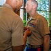 Two U.S. Marine Corps personnel in tan service uniform (Charlie uniform) are engaged in a military decoration ceremony indoors. A senior Marine is pinning a medal or award onto the chest of another Marine who is wearing multiple service ribbons. The setting appears to be a formal indoor venue with large windows and greenery visible in the background.