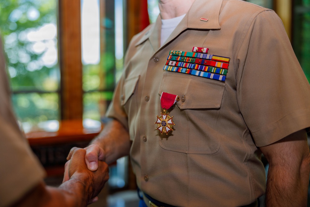 A U.S. Marine Corps service member in tan/khaki uniform is shaking hands during what appears to be a formal military ceremony. The individual is wearing a Legion of Merit medal along with multiple rows of service ribbons on their chest. The setting appears to be an indoor venue with wooden furnishings and natural light from windows.