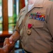 A U.S. Marine Corps service member in tan/khaki uniform is shaking hands during what appears to be a formal military ceremony. The individual is wearing a Legion of Merit medal along with multiple rows of service ribbons on their chest. The setting appears to be an indoor venue with wooden furnishings and natural light from windows.