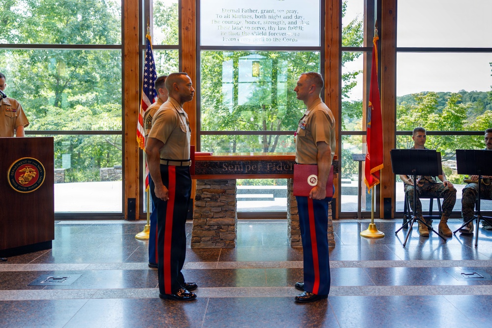 A formal United States Marine Corps ceremony is taking place in an indoor venue with large windows overlooking a wooded area. Two Marines in dress blues with red stripe trousers stand facing each other at a stone podium inscribed with 'Semper Fidelis,' with one holding a red folder bearing the Marine Corps emblem. Additional personnel including musicians and a speaker at a podium are visible in the background.