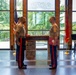 A formal United States Marine Corps ceremony is taking place in an indoor venue with large windows overlooking a wooded area. Two Marines in dress blues with red stripe trousers stand facing each other at a stone podium inscribed with 'Semper Fidelis,' with one holding a red folder bearing the Marine Corps emblem. Additional personnel including musicians and a speaker at a podium are visible in the background.