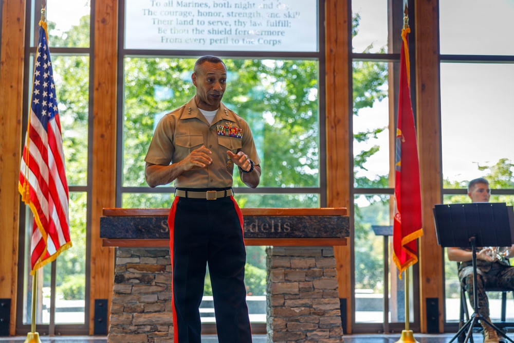 A U.S. Marine Corps officer in service uniform is speaking at a podium during what appears to be an official military ceremony or event. The setting includes an American flag on the left and a Marine Corps flag on the right, with a text inscription visible on the window behind him. A musician is partially visible in the background to the right.
