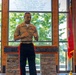 A U.S. Marine Corps officer in service uniform is speaking at a podium during what appears to be an official military ceremony or event. The setting includes an American flag on the left and a Marine Corps flag on the right, with a text inscription visible on the window behind him. A musician is partially visible in the background to the right.
