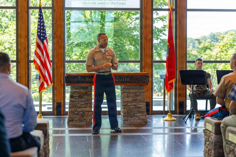 A U.S. Marine Corps officer in dress uniform is delivering a speech or address at what appears to be a formal military ceremony. The setting features a stone podium with the inscription 'Semper Fidelis,' flanked by the U.S. flag and a Marine Corps flag. An audience is seated facing the speaker, with another Marine visible in the background.