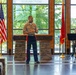 A U.S. Marine Corps officer in dress uniform is delivering a speech or address at what appears to be a formal military ceremony. The setting features a stone podium with the inscription 'Semper Fidelis,' flanked by the U.S. flag and a Marine Corps flag. An audience is seated facing the speaker, with another Marine visible in the background.