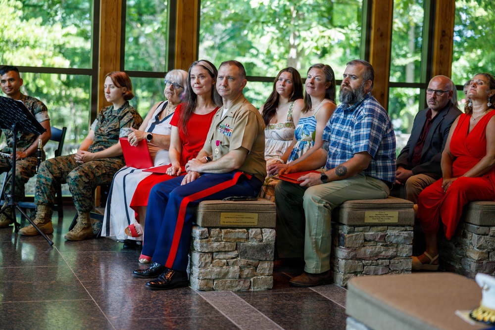 A formal military ceremony is taking place in an indoor pavilion-style venue surrounded by trees. A U.S. Marine Corps officer in dress uniform with medals is seated alongside civilians, likely family members, suggesting a retirement or awards ceremony. The attendees are dressed formally, with several wearing red, and are attentively watching a presentation off-camera.