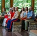 A formal military ceremony is taking place in an indoor pavilion-style venue surrounded by trees. A U.S. Marine Corps officer in dress uniform with medals is seated alongside civilians, likely family members, suggesting a retirement or awards ceremony. The attendees are dressed formally, with several wearing red, and are attentively watching a presentation off-camera.