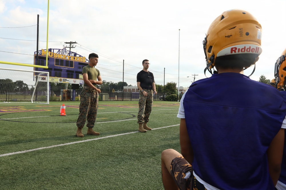 Two individuals in U.S. Marine Corps camouflage uniforms are standing on a turf athletic field, appearing to address or instruct a group of football players. The setting is a high school football field belonging to the 'Musketeers' team, visible from the scoreboard/sign in the background. A football player wearing a gold Riddell helmet and purple jersey is visible in the foreground, seated and facing the military personnel.