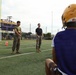 Two individuals in U.S. Marine Corps camouflage uniforms are standing on a turf athletic field, appearing to address or instruct a group of football players. The setting is a high school football field belonging to the 'Musketeers' team, visible from the scoreboard/sign in the background. A football player wearing a gold Riddell helmet and purple jersey is visible in the foreground, seated and facing the military personnel.