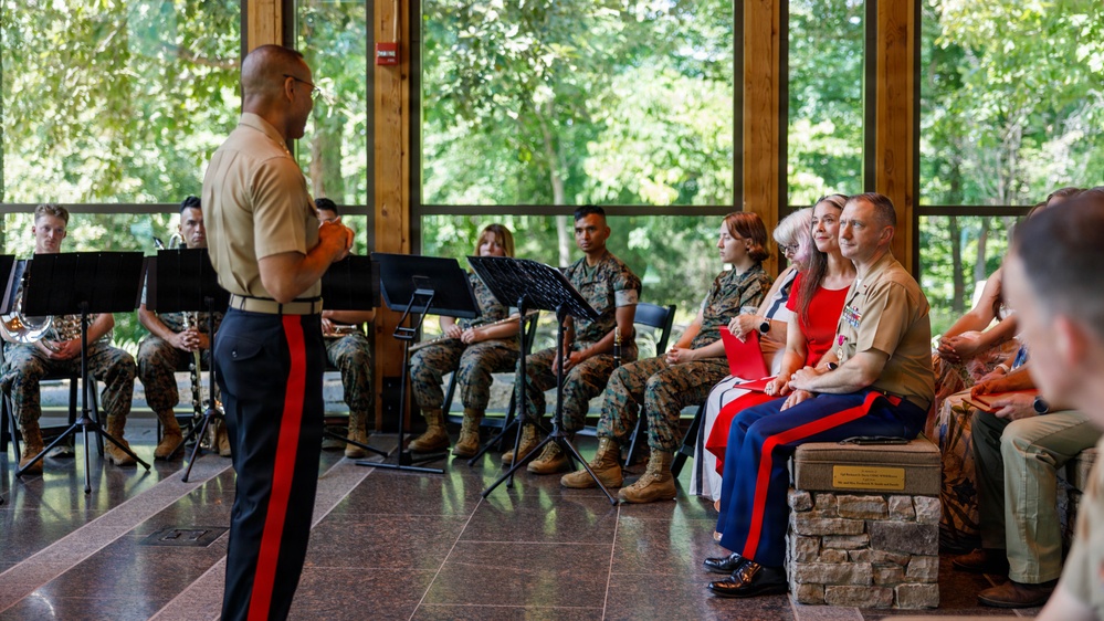 A U.S. Marine Corps officer in dress uniform stands at the front of an indoor pavilion addressing a seated audience of fellow Marines and civilians. A military band with music stands and instruments is visible to the left, suggesting a formal ceremony or performance event. The setting appears to be a modern glass-walled venue surrounded by lush green trees.