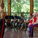 A U.S. Marine Corps officer in dress uniform stands at the front of an indoor pavilion addressing a seated audience of fellow Marines and civilians. A military band with music stands and instruments is visible to the left, suggesting a formal ceremony or performance event. The setting appears to be a modern glass-walled venue surrounded by lush green trees.