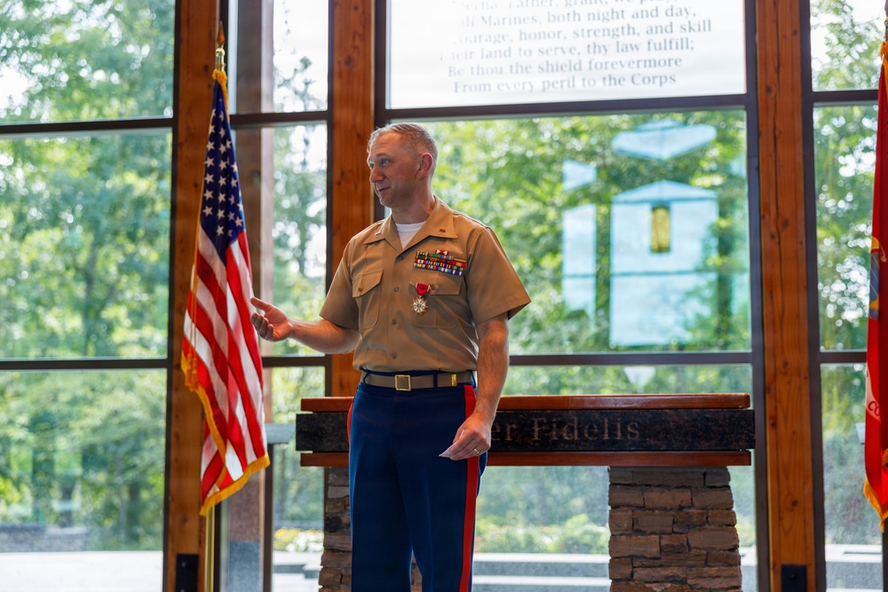 A U.S. Marine Corps officer in service uniform (Charlie uniform) stands at a podium bearing the inscription 'Semper Fidelis' and delivers a speech or remarks at a formal ceremony. He is wearing multiple service ribbons and a medal, consistent with a retirement or award ceremony. The setting appears to be a Marine Corps chapel or ceremonial hall with large windows overlooking trees.