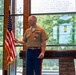 A U.S. Marine Corps officer in service uniform (Charlie uniform) stands at a podium bearing the inscription 'Semper Fidelis' and delivers a speech or remarks at a formal ceremony. He is wearing multiple service ribbons and a medal, consistent with a retirement or award ceremony. The setting appears to be a Marine Corps chapel or ceremonial hall with large windows overlooking trees.