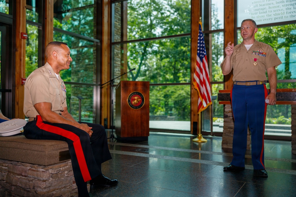 Two U.S. Marine Corps officers are participating in what appears to be a formal military ceremony or award presentation in an indoor venue. One Marine is seated and listening while the other stands and speaks, gesturing with one hand. The standing Marine is wearing a medal, suggesting this may be an awards or retirement ceremony.