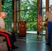 Two U.S. Marine Corps officers are participating in what appears to be a formal military ceremony or award presentation in an indoor venue. One Marine is seated and listening while the other stands and speaks, gesturing with one hand. The standing Marine is wearing a medal, suggesting this may be an awards or retirement ceremony.