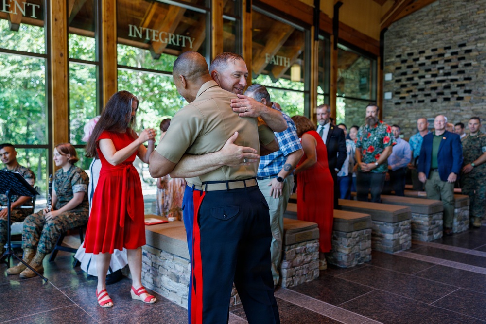 A military ceremony taking place in a modern pavilion or chapel-like venue with words such as 'INTEGRITY' and 'FAITH' displayed on the glass walls. A U.S. Marine in dress uniform (tan shirt, blue trousers with red stripe) is embracing another individual in a warm hug, suggesting a retirement, promotion, or change-of-command ceremony. Attendees including other Marines in uniform and civilians in formal attire look on from the background.