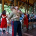 A military ceremony taking place in a modern pavilion or chapel-like venue with words such as 'INTEGRITY' and 'FAITH' displayed on the glass walls. A U.S. Marine in dress uniform (tan shirt, blue trousers with red stripe) is embracing another individual in a warm hug, suggesting a retirement, promotion, or change-of-command ceremony. Attendees including other Marines in uniform and civilians in formal attire look on from the background.