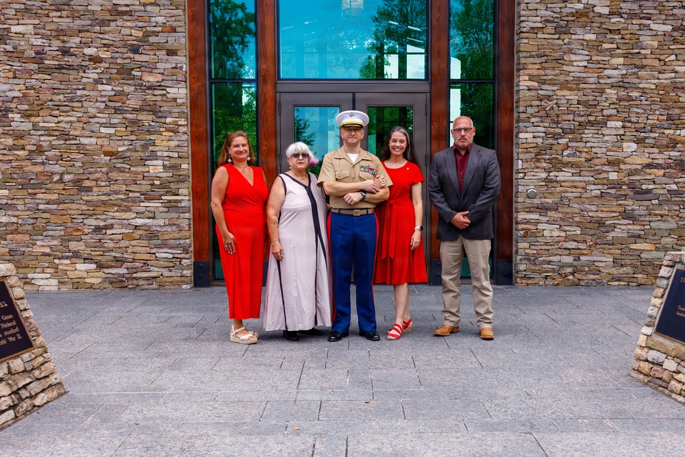 A group of five people pose for a formal photograph in front of a modern stone-and-glass building entrance. The central figure is a U.S. Marine Corps officer in dress uniform, flanked by four civilians dressed in semi-formal attire. The setting appears to be a memorial or civic building, suggested by the stone plaques visible on either side of the frame.
