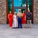 A group of five people pose for a formal photograph in front of a modern stone-and-glass building entrance. The central figure is a U.S. Marine Corps officer in dress uniform, flanked by four civilians dressed in semi-formal attire. The setting appears to be a memorial or civic building, suggested by the stone plaques visible on either side of the frame.