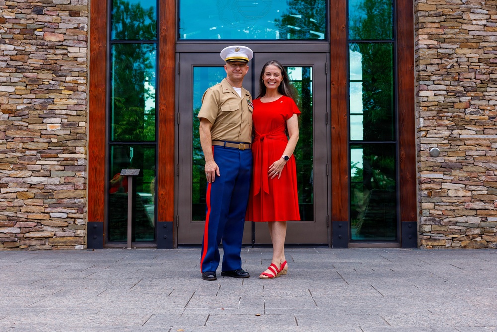 A man in U.S. Marine Corps Service 'C' uniform (khaki shirt, blue trousers with red blood stripe, white cover) stands with a woman in a red dress in front of a modern stone-and-glass building entrance. Both are smiling and posing together, suggesting a formal or celebratory occasion. The setting appears to be an outdoor plaza or entrance area.