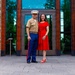 A man in U.S. Marine Corps Service 'C' uniform (khaki shirt, blue trousers with red blood stripe, white cover) stands with a woman in a red dress in front of a modern stone-and-glass building entrance. Both are smiling and posing together, suggesting a formal or celebratory occasion. The setting appears to be an outdoor plaza or entrance area.