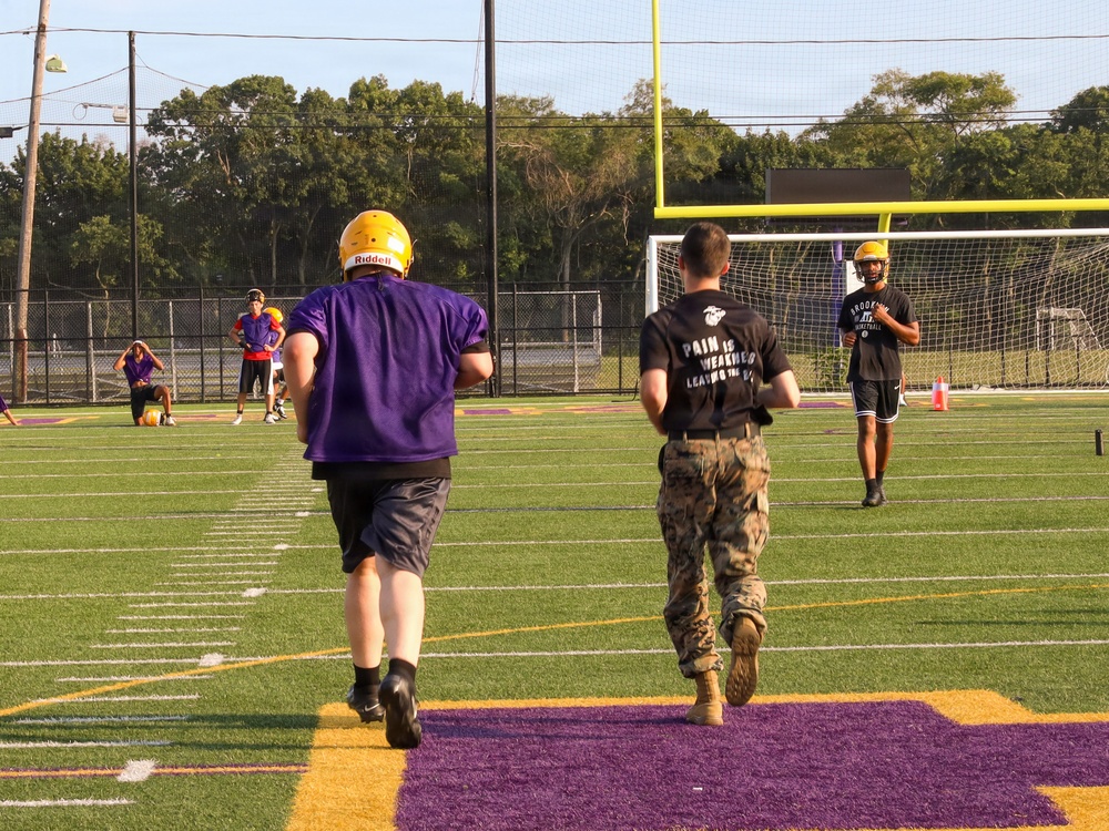 A football practice or training session is taking place on a purple and gold turf field. Players in purple jerseys and gold Riddell helmets are running drills alongside individuals in military camouflage uniforms and black t-shirts with the text 'Pain is Weakness Leaving the Body.' The scene suggests a military-meets-athletics training event, possibly a recruiting or community engagement activity.