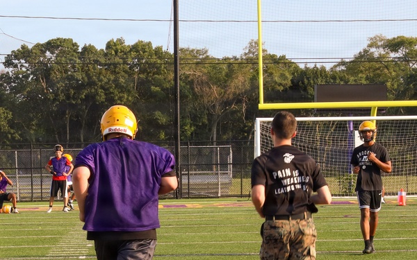A football practice or training session is taking place on a purple and gold turf field. Players in purple jerseys and gold Riddell helmets are running drills alongside individuals in military camouflage uniforms and black t-shirts with the text 'Pain is Weakness Leaving the Body.' The scene suggests a military-meets-athletics training event, possibly a recruiting or community engagement activity.