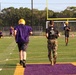 A football practice or training session is taking place on a purple and gold turf field. Players in purple jerseys and gold Riddell helmets are running drills alongside individuals in military camouflage uniforms and black t-shirts with the text 'Pain is Weakness Leaving the Body.' The scene suggests a military-meets-athletics training event, possibly a recruiting or community engagement activity.