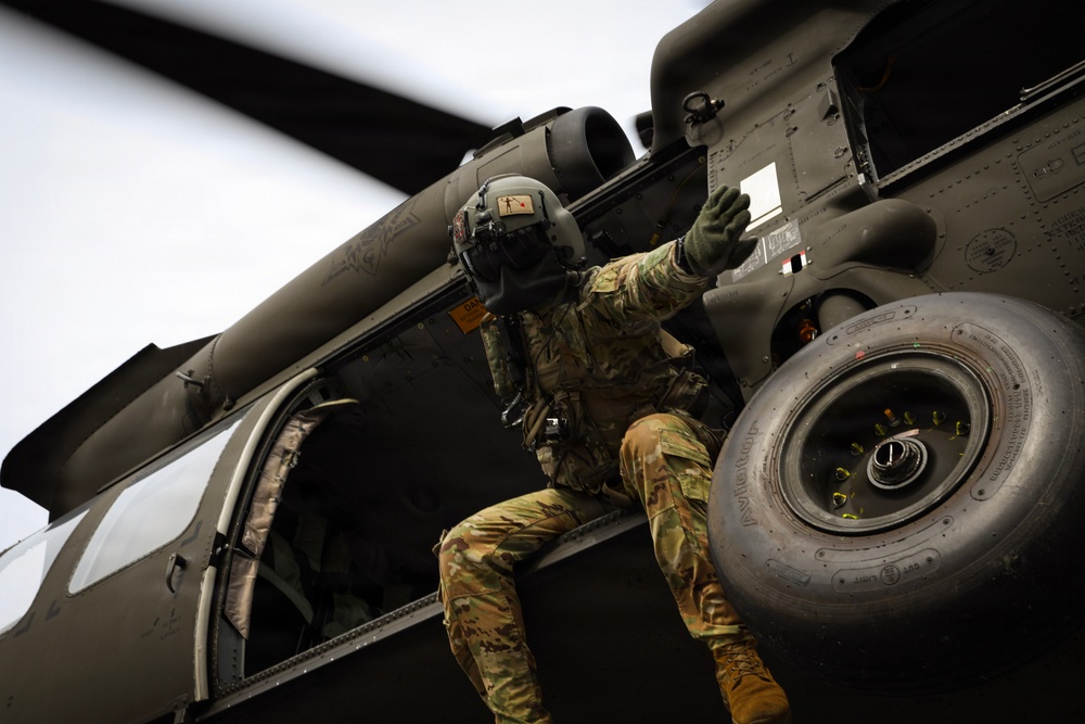 A military aviator or crew member in full combat gear and flight helmet is climbing onto or inspecting the exterior of a military helicopter, likely a UH-60 Black Hawk. The individual is wearing multicam/OCP camouflage uniform and appears to be conducting a pre-flight or post-flight inspection. The image is taken from a low angle, emphasizing the scale of the aircraft and the crew member's activity.